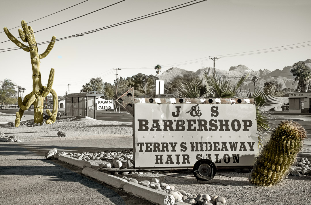 Sign for J&S Barbershop surrounded by cactus. Along side road and small mountains.