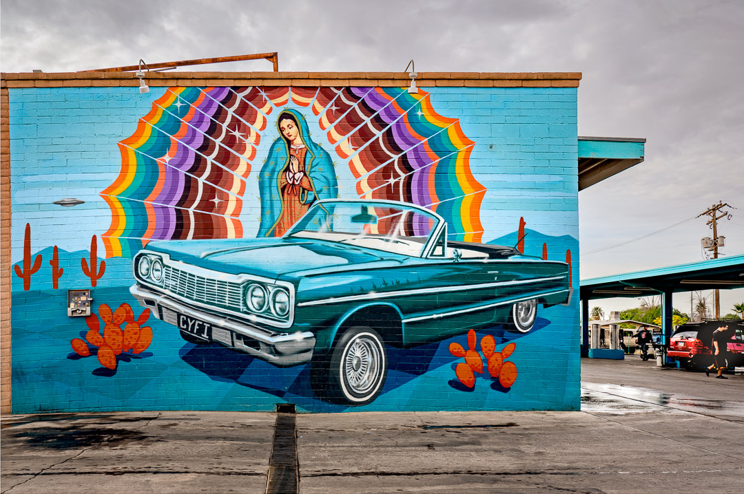 Mural of lowrider car on brick wall with Virgin Mary, cactus, and UFO. Car wash in background. South Tuscon.