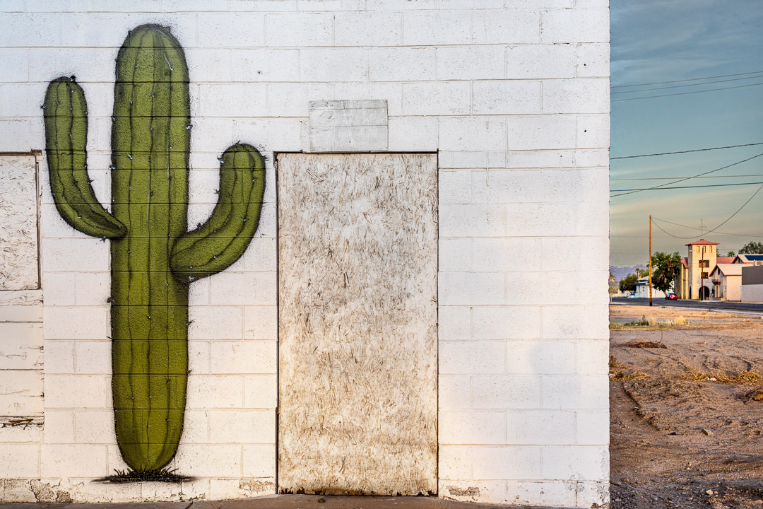 Saguaro mural on white brick wall. Dirt lot on right side. Twentynine Palms, CA