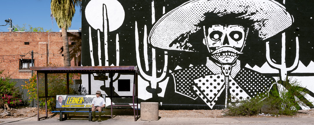 Panoramic image of B&W mural of skeleton cowboy in front of saguaro cactus and moon. Mural is behind man sitting at bus stop. Tuscon.
